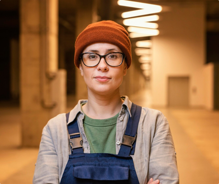 Professional woman with glasses and orange beanie wearing overalls in a workshop environment